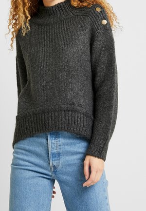 Kvinde iført en mørkegrå strikket sweater med to metalliske knapper på skulderen og lyseblå jeans mod en ensfarvet baggrund.