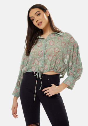 Light green, floral-patterned cropped blouse with a button-up front, collar, and gathered sleeves, featuring a tie closure at the waist.