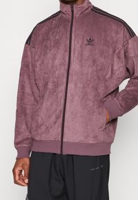 Lila velour zip-up jacka med hög krage och svarta tre-randiga detaljer på axlarna, med en broderad Adidas-logotyp på bröstet.