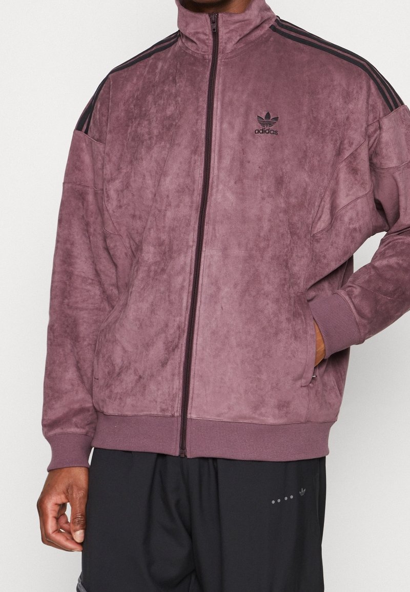 Lila velour zip-up jacka med hög krage och svarta tre-randiga detaljer på axlarna, med en broderad Adidas-logotyp på bröstet.