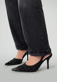 Escarpins noirs avec une bride arrière en tissu texturé et un bout en verni noir brillant. Ils ont un bout pointu et un talon aiguille fin.