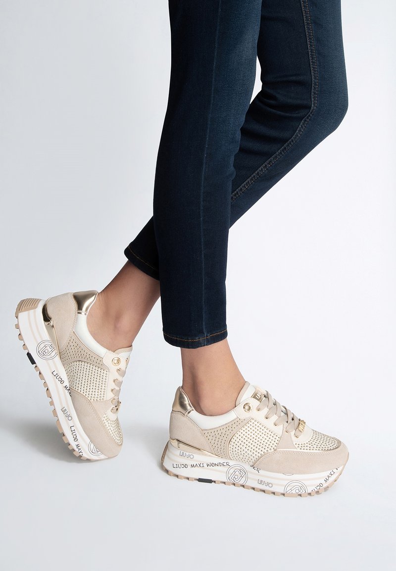 Scarpe sportive beige con pannelli in rete e suede, dettagli metallici, design con lacci e suole appositamente decorate, indossate con jeans scuri.