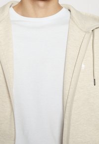 Närbild av en person som bär en enfärgad vit T-shirt med rund hals under en oöppen ljusbeige zip-up hoodie med dragsnören.