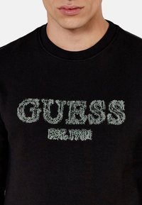 Felpa nera con logo "GUESS" in dettagliata cucitura verde, con "EST. 1981" sottostante. Collo a giro facile e morbido al tatto.