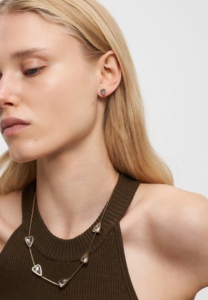 Blond kvinde med hjerteformede øreringe og matchende halskæde iført en mørk ærmeløs ribbet top, vender til venstre mod en ensfarvet baggrund.