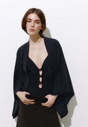 Un mannequin pose devant un mur blanc, vêtu d’une robe noire et d’un châle ample, en tenant une pochette noire.