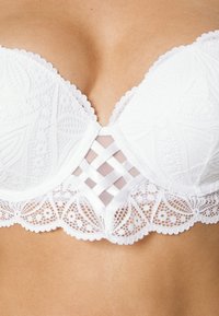 Soutien-gorge en dentelle blanche avec des motifs floraux, bonnets texturés et un panneau en mesh transparent présentant des détails en croix au centre. Bords festonnés.