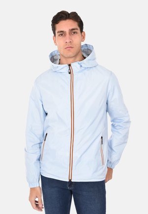 Giovane uomo con giacca con cappuccio azzurro chiaro, con cerniera a righe marroni e tasche con zip, in piedi davanti a uno sfondo semplice.