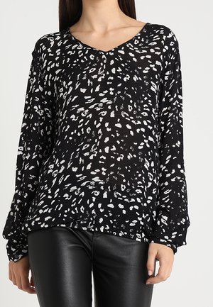 Czarna bluzka z długim rękawem w białe abstrakcyjne plamy, zestawiona z czarnymi skórzanymi spodniami na stojącej osobie.