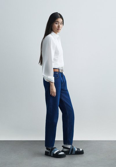 Egy nő oldalra állva fehér inget, kék farmert, zoknival viselt szandált és jól látható MOSCHINO logóval ellátott övet visel.