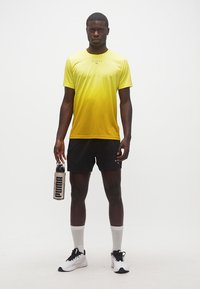 Άνδρας που φοράει κίτρινη μπλούζα με διαβάθμιση χρωμάτων, μαύρη σορτς, λευκές κάλτσες και αθλητικά παπούτσια, κρατώντας ένα παγούρι PUMA, στέκεται μπροστά σε λευκό φόντο.