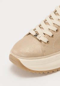 Baskets beiges avec une tige brillante et texturée, dotées de lacets crème et d'accents dorés sur les œillets et le logo, avec une semelle en caoutchouc épaisse.