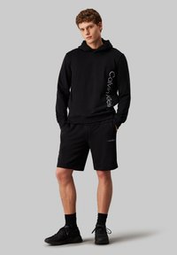 Felpa nera con cappuccio e logo bianco sul lato, abbinata a pantaloni corti neri con un piccolo logo. Entrambi sono realizzati in materiale morbido.
