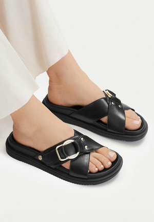 Füße tragen schwarze Slide-Sandalen mit gekreuzten Riemen und silbernen Schnallendetails auf weißem Hintergrund.