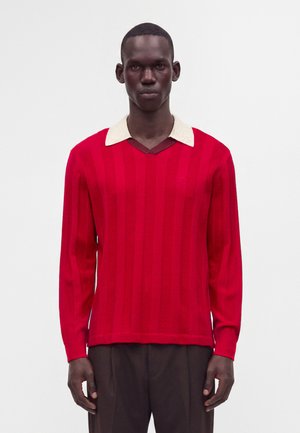 Homme portant une chemise rouge à manches longues avec des rayures verticales et un col blanc, associée à un pantalon marron foncé, se tenant devant un fond uni.
