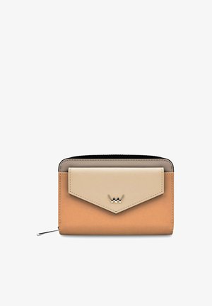 Hellgrünes, geripptes Oberteil, weiße Hosen mit weitem Bein und weiße Turnschuhe. Hält eine kleine beigefarbene Brieftasche. Umgeben von Topfpflanzen in einem hellen Raum.