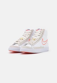 Un paio di sneakers high-top Nike bianche e beige con il logo swoosh arancione e lacci bianchi su uno sfondo neutro.