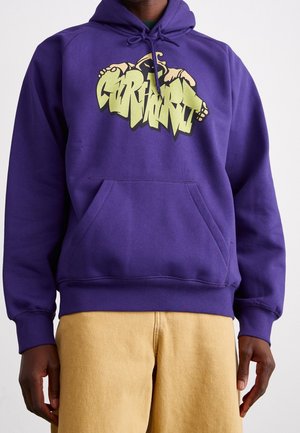 Person wearing a purple hoodie with graffiti-style text graphic and beige pants, standing against a plain light background.