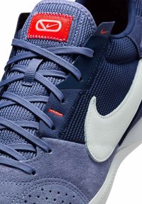 Zili zamšas un acs kedas ar baltu Nike simbolu, sarkaniem akcentiem un pelēkām auklēm. Rakstainais audums ir dažādās daļās.