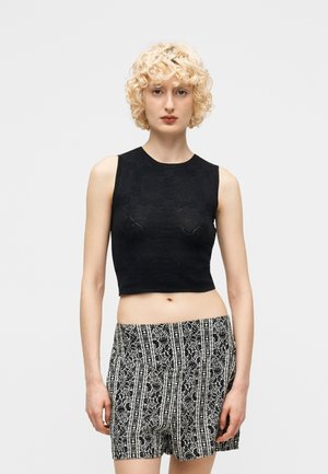 Blonde Person mit lockigem Haar, die ein ärmelloses schwarzes Croptop und hoch taillierte schwarz-weiße gemusterte Shorts vor einem schlichten Hintergrund trägt.