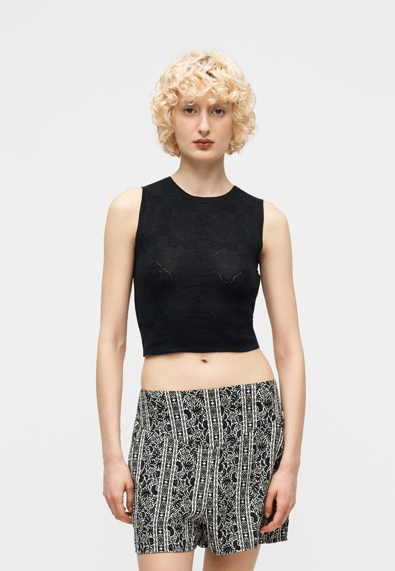 Ξανθό άτομο με σγουρά μαλλιά που φοράει μαύρο crop top χωρίς μανίκια και ψηλόμεσα σορτς με ασπρόμαυρο μοτίβο, μπροστά από απλό φόντο.