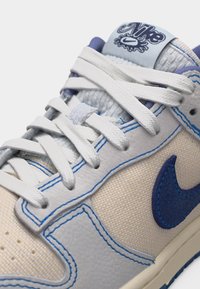 Zapatilla Nike blanca y beige con costuras azules, logo Swoosh azul, cordones blancos y lengüeta gris con logotipo de Nike estilo graffiti.