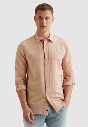 Homme portant une chemise rose clair boutonnée avec les manches retroussées et un jean bleu clair, debout avec une main dans la poche.
