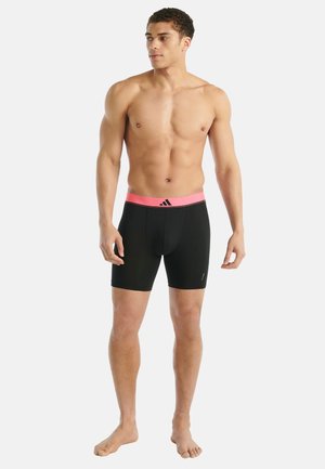 Oberkörperfreier Mann trägt schwarze sportliche Boxershorts mit einem rosa Bund und Adidas-Logo, steht barfuß vor einem weißen Hintergrund.