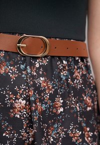 Ceinture en cuir marron avec une boucle ovale dorée, portée sur une jupe noire à fleurs avec de petites fleurs roses et bleues sur un fond sombre.