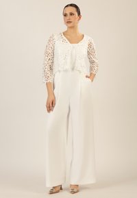 Weißer Jumpsuit mit weitem Bein, ausgestattet mit einer transparenten Spitzenbolero mit floralen Mustern und drei Viertel Ärmeln. Silberne High Heels runden den Look ab.