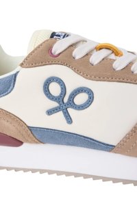 Zapatilla de deporte en tonos beige, marrón y azul con cordones blancos y un logo de tijeras bordado en el lateral, con suela blanca y fondo estriado.