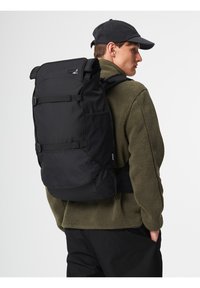 Black backpack with roll-top closure, adjustable straps, and side pockets, worn by a person in a green fleece jacket. Durable material, minimalist design.