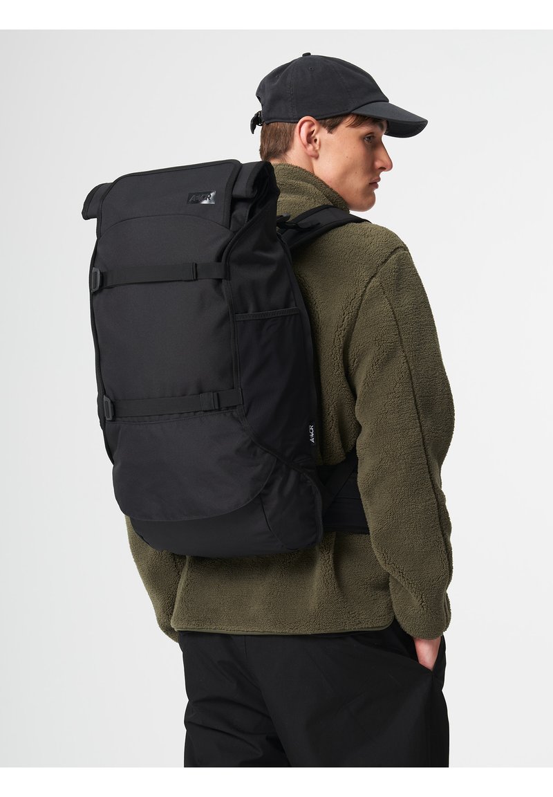 Black backpack with roll-top closure, adjustable straps, and side pockets, worn by a person in a green fleece jacket. Durable material, minimalist design.