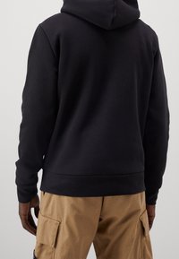 Felpa nera con cappuccio, tasca a marsupio frontale, polsini e orlo a costine. Realizzata in materiale morbido, indossata con pantaloni cargo marrone chiaro.