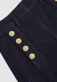Mørkeblå denimstof med fire dekorative guldknapper arrangeret lodret nær en bæltestrop.