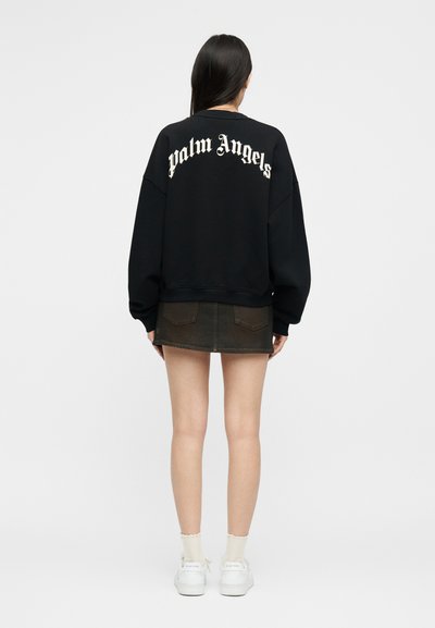 Γυναίκα που στέκεται με το σώμα στραμμένο προς τα πίσω, φορώντας μαύρο φούτερ με το κείμενο "palm angels", σκούρα μίνι φούστα, λευκές κάλτσες και λευκά αθλητικά παπούτσια.