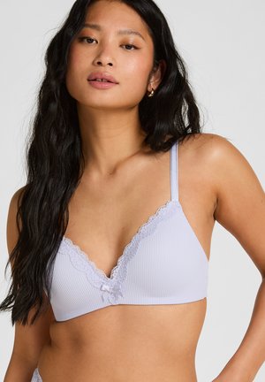 Frau mit langen schwarzen Haaren, die einen hellvioletten BH mit Spitzenbesatz und dünnen Trägern vor einem schlichten hellen Hintergrund trägt.