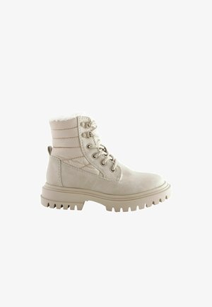 Beige nilkkuri, jossa on teksturoitu päällinen, pehmustettu kaulus ja nauhakiinnitys edessä. Varustettu karkealla pohjalla, jossa on voimakkaat urat pidon parantamiseksi.