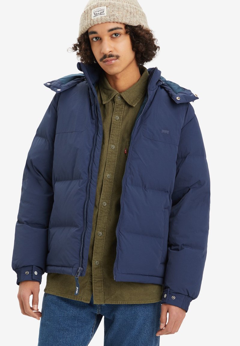 Chaqueta acolchada azul marino con cremallera en la parte delantera, capucha desmontable y puños elásticos, llevada sobre una camisa de pana verde oliva. El tejido es suave y mate.