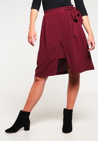 Jupe portefeuille bordeaux avec une ceinture nouée et une fente à l'avant. Fabriquée en tissu texturé, associée à des bottines noires.