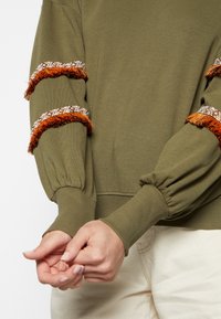 Maglione verde oliva con maniche a sbuffo, caratterizzato da frange intrecciate arancioni e bianche lungo le maniche. Dettagli a coste ai polsini e all'orlo.