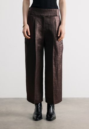 Mujer con pantalones anchos recortados de color marrón oscuro brillante, botines negros, top negro sin mangas y una pulsera dorada en la muñeca izquierda.