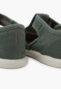 Grüne Canvas-Slipper mit strukturiertem Obermaterial, weißer Gummisohle und sichtbaren Nähten. Offene Seiten für Atmungsaktivität.