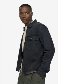Dark denim jacket with a relaxed fit, collar, and a front pocket. Features a full zipper, textured fabric, and long sleeves.