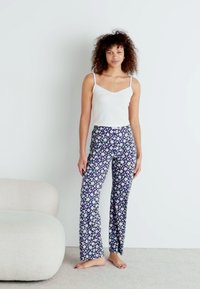 Λευκό crop top, συνδυασμένο με μπλε patterned παντελόνι με λουλουδάτα σχέδια. Το μοντέλο στέκεται ξυπόλητο μπροστά σε έναν ανοιχτό τοίχο και στον απαλό καναπέ.