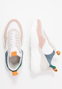 Baskets blanches avec des empiècements en daim beige et bleu, semelles blanches épaisses, languettes orange et lacets blancs, vues de dessus et de côté.