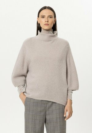Woman wearing a light gray high-neck sweater with three-quarter sleeves and gray plaid pants, standing with one hand in her pocket.