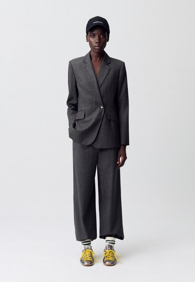 Persona vistiendo un traje oversized gris oscuro, gorra negra, calcetines blancos con rayas verdes y zapatillas metálicas con cordones amarillos, de pie contra un fondo simple.