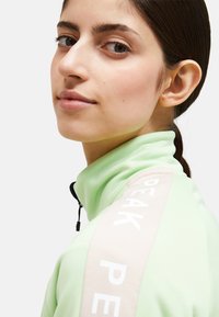 Giacca atletica verde chiaro con zip e colletto alto, dettagli in nero e un accento del logo beige sulla spalla. Tessuto liscio.