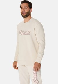 Sweat-shirt de couleur crème avec le logo "asics" rouge, col rond, manches longues et poignets côtelés. Texture lisse avec une coupe décontractée.